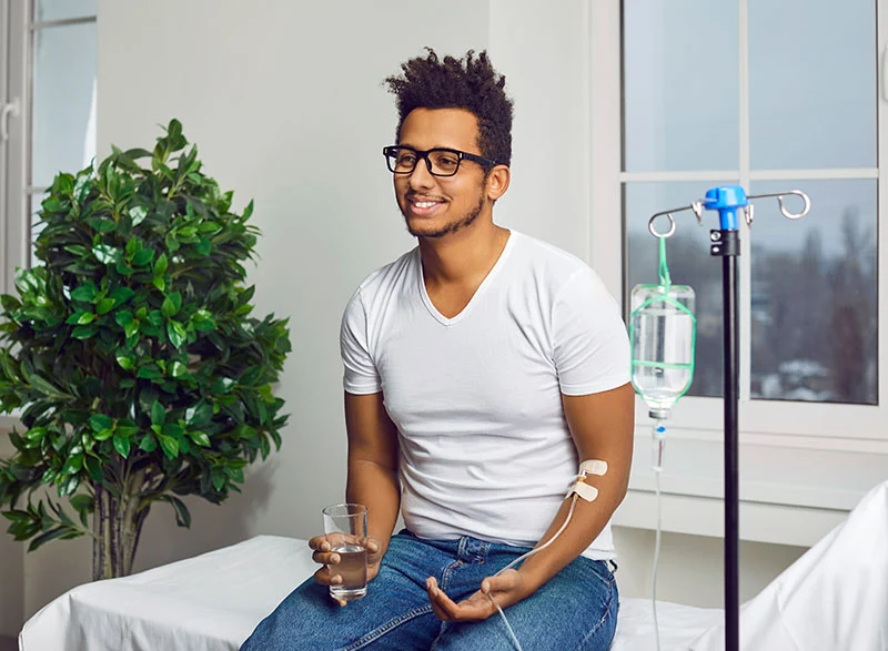 A smiling man sits on an examination table with an IV drip in his arm, holding a glass of water. A smiling man sits on an examination table with an IV drip in his arm, holding a glass of water.